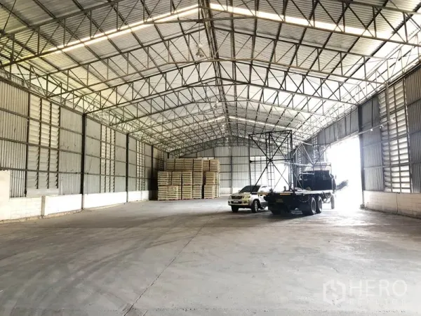 Warehouse for rent in Pathum Thani - Spacious warehouse interior with an 8m steel truss roof, concrete floor, and parked trucks.