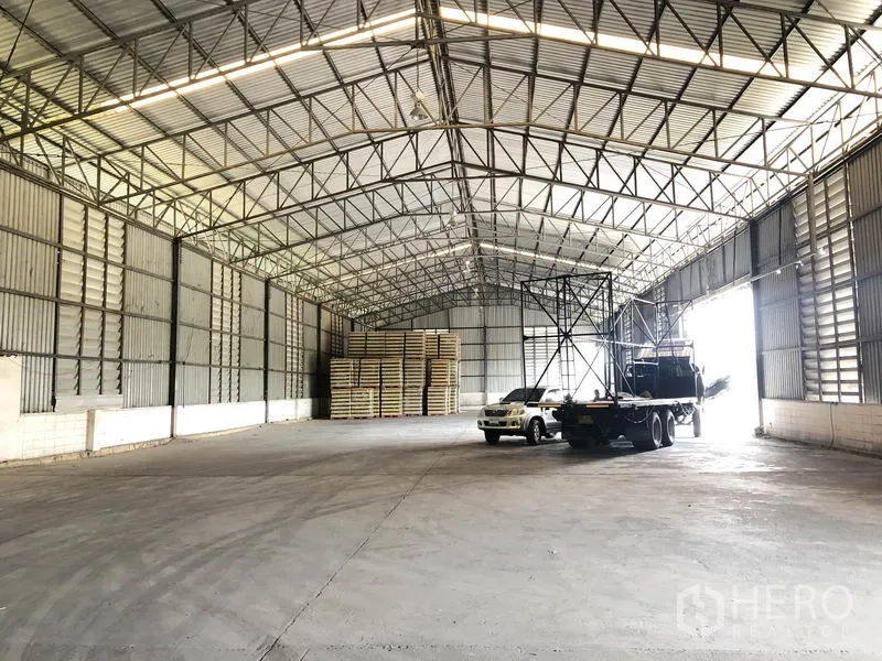 Warehouse for rent in Pathum Thani - Spacious warehouse interior with an 8m steel truss roof, concrete floor, and parked trucks.