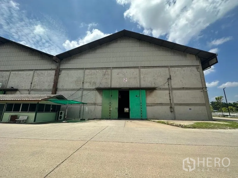 Warehouse for rent in Mueang Pathum Thani, Pathum Thani - Front facade of a warehouse bay with large green sliding door, loading apron and small guard booth.