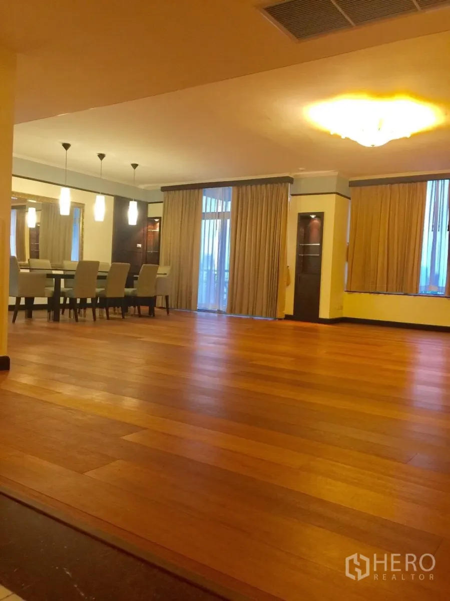 Condominium for rent in Pathum Wan, Bangkok - Spacious open-plan living and dining area with warm wood floors, pendant lights and floor-to-ceiling curtains opening to a balcony.