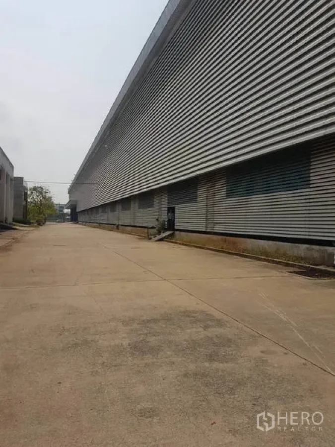 Factory for rent in Mueang Samut Sakhon, Samut Sakhon - Wide concrete driveway beside a long corrugated‑metal factory exterior with multiple loading bays in Samut Sakhon.