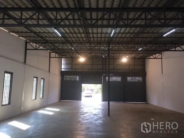 Warehouse for rent in Pathum Thani - Spacious warehouse interior featuring a high steel-truss ceiling, concrete floor, and large roller shutter entry for vehicle access.