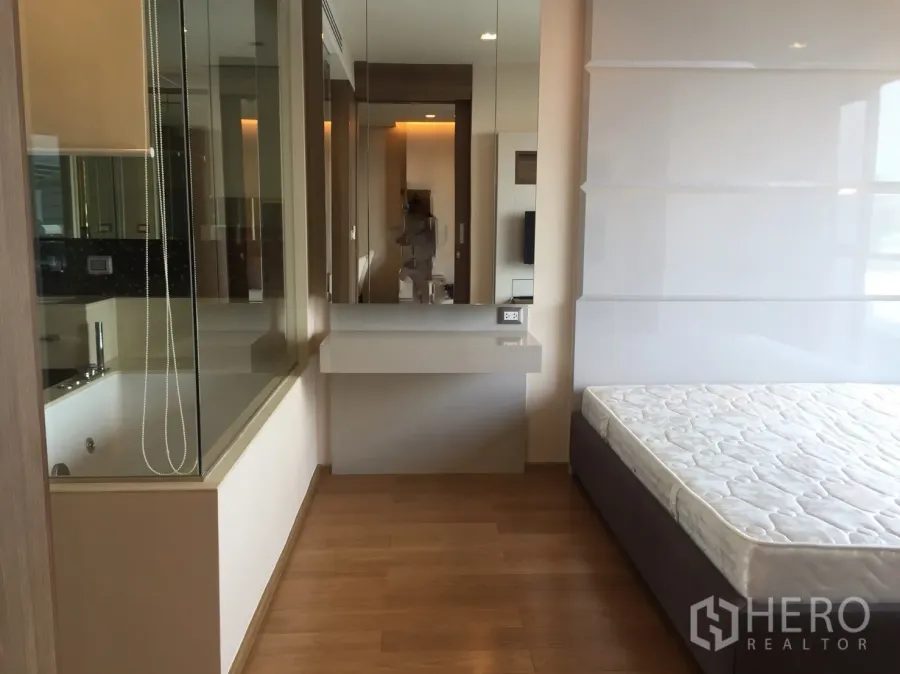 Condominium for rent in Bang Rak, Bangkok - Bright bedroom with wood flooring, a vanity desk and a glass partition looking into a bathroom with a soaking tub.