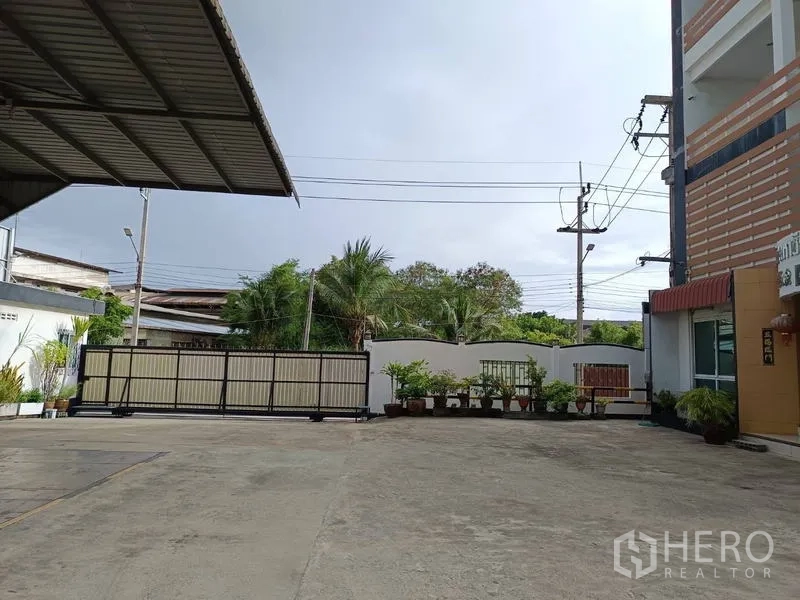Factory for sale in Ban Phaeo, Samut Sakhon - Secure sliding gate opening to spacious concrete forecourt with power lines and greenery.