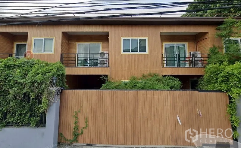 联排别墅 出租或出售在Watthana, Bangkok - 两层现代联排别墅外观，木质立面、双阳台、推拉玻璃门与黑色栏杆，前方有围墙与绿篱，位于素坤逸—Ekkamai片区。