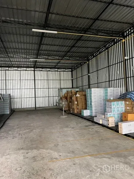 Warehouse for rent in Lat Phrao, Bangkok - Spacious aisle inside a metal-roof warehouse with concrete flooring, long strip lighting, pallets of boxes, and a standing fan.