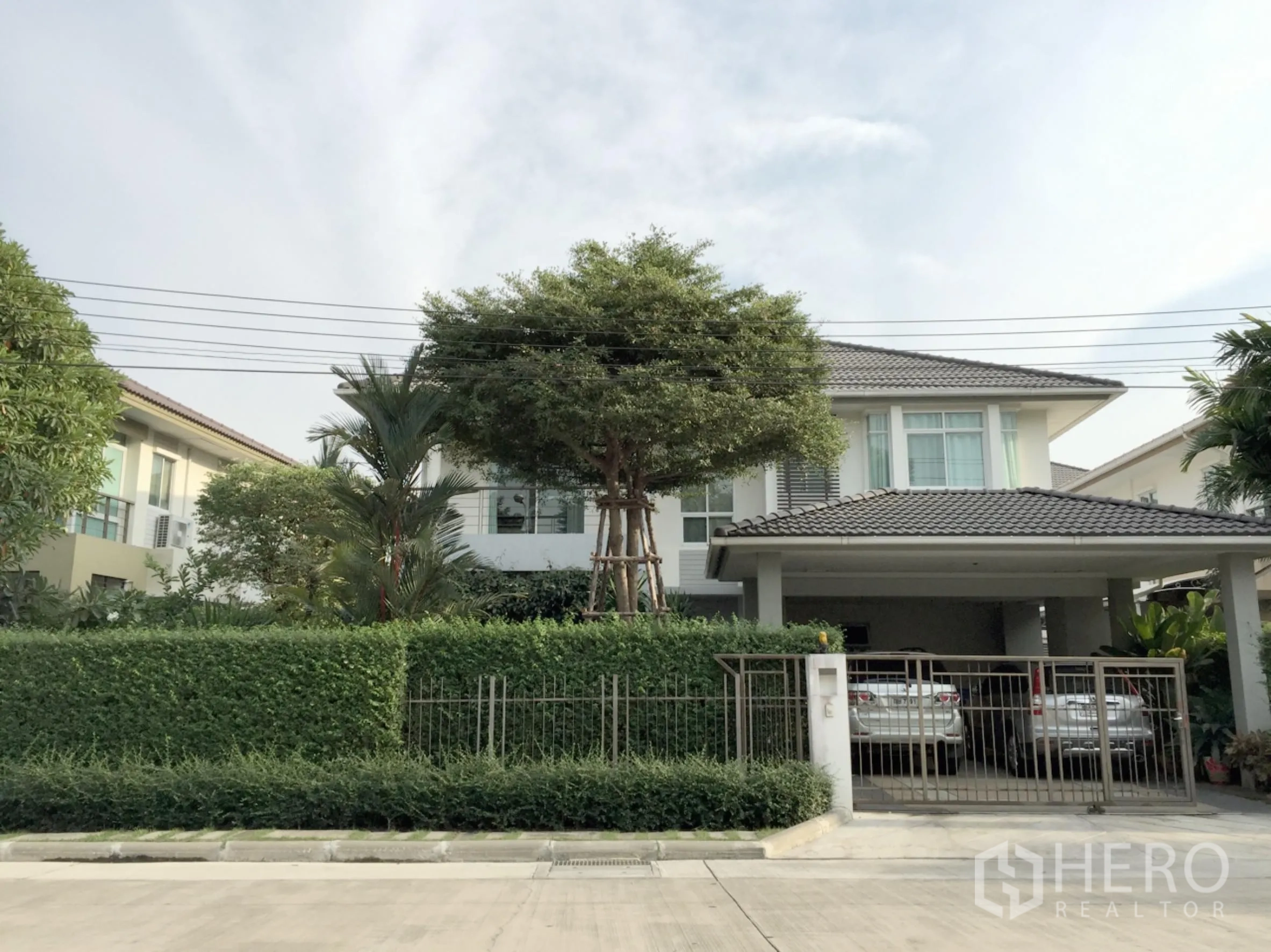 Detached House for sale in Khan Na Yao, Bangkok - Two-storey detached house with lush hedge, large tree, gated entrance and covered two-car carport in a quiet Khan Na Yao neighborhood.