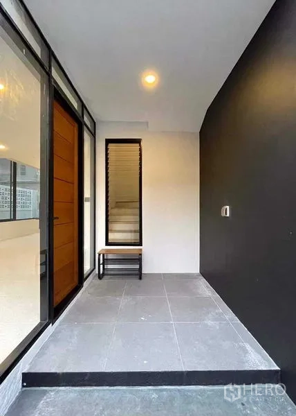 Home Office for sale in Lat Phrao, Bangkok - Covered front porch with a bench beside full-height glass and wood door.
