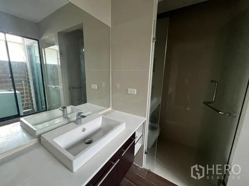 Townhouse for rent or sale in Khlong Toei, Bangkok - Bathroom vanity with a wide mirror beside a glass-enclosed shower and window.