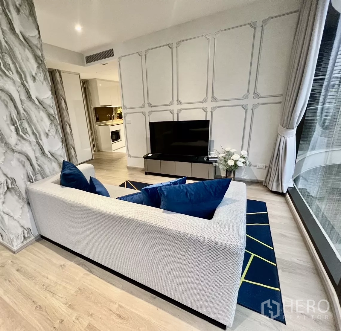 Condominium for rent in Khlong Toei, Bangkok - Living room with sofa and TV console by a large window; kitchen visible beyond.
