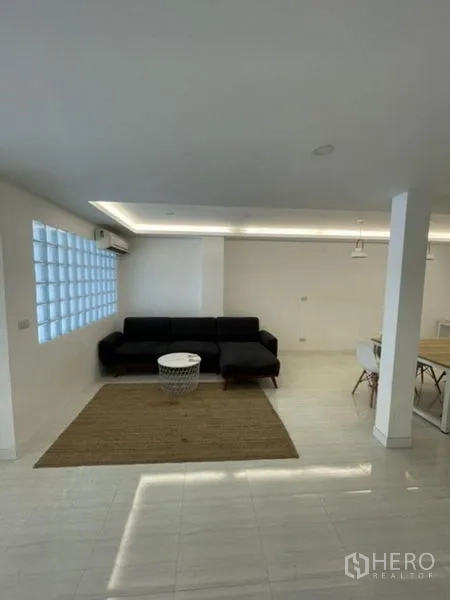 Townhouse for rent in Phra Khanong, Bangkok - Open-plan living area with black sofa, recessed lighting and a glass-block window.