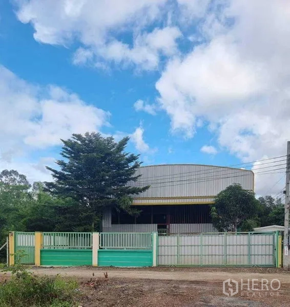 Warehouse for rent in Lat Krabang, Bangkok - Gated steel warehouse exterior with wide driveway and trees under a blue sky in Lat Krabang.