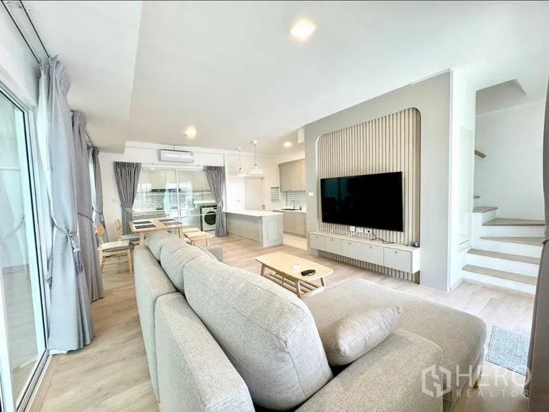Townhouse for rent or sale in Prawet, Bangkok - Cozy living room with sectional sofa facing a large TV, open to kitchen and stairs.