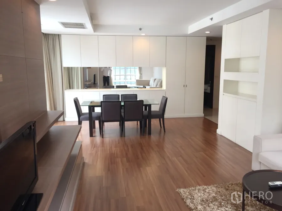 Condominium for rent in Pathum Wan, Bangkok - Bright open-plan living and dining area with wood flooring, glass dining table and built-in white cabinetry with a large mirror.