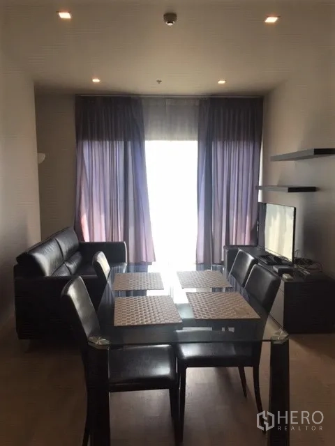 Condominium for rent in Bangkok - Open-plan living and dining area with glass table, black leather sofa, TV console and floor-to-ceiling curtains in a 2-bedroom condo near BTS Phrom Phong.