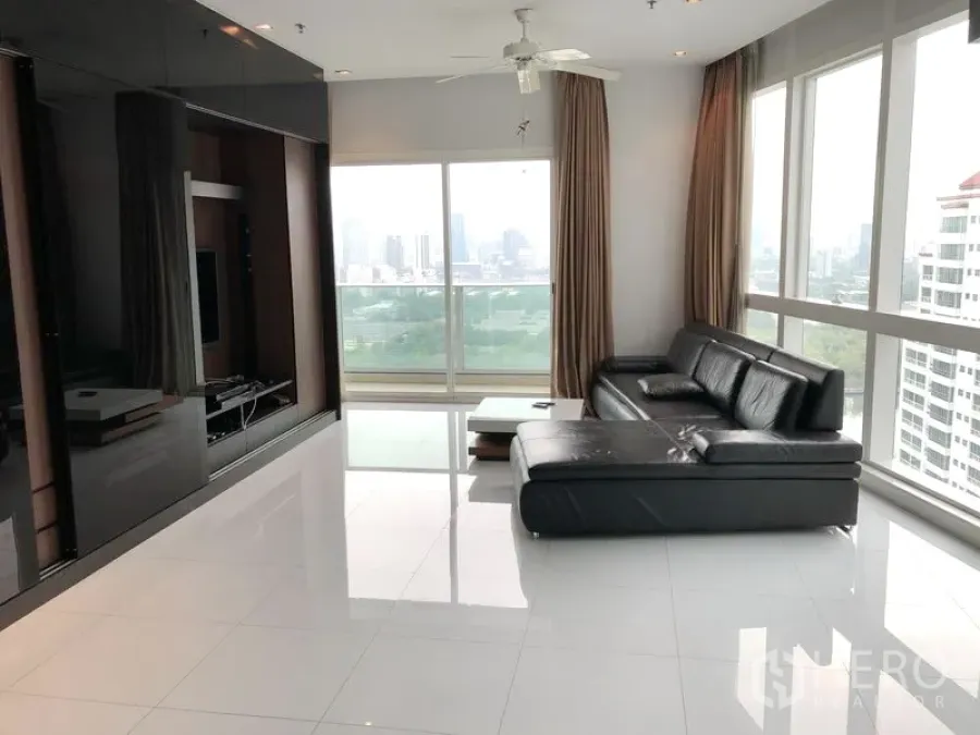 Condominium for rent in Khlong Toei, Bangkok - High-floor living room with black leather sectional, glossy white tiles, and floor-to-ceiling windows showing park and skyline views.