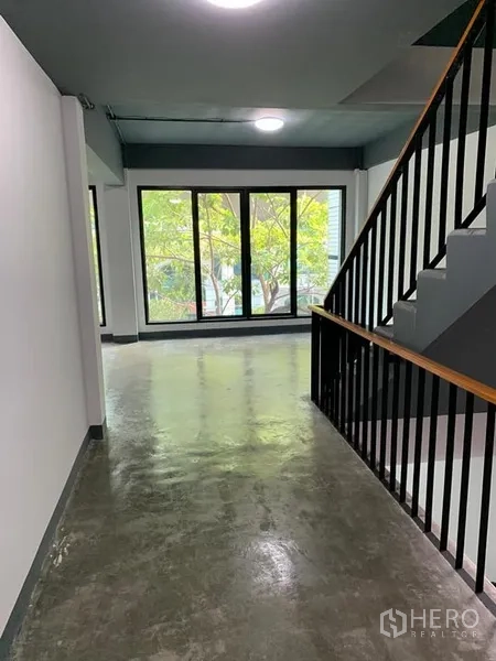 Shophouse for rent in Khlong Toei, Bangkok - Corridor with black metal staircase and floor-to-ceiling windows overlooking greenery.