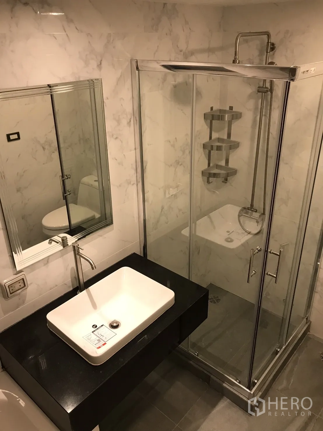 Condominium for rent or sale in Khlong Toei, Bangkok - Bathroom with glass-enclosed shower, square basin on black vanity and marble-style wall tiles.