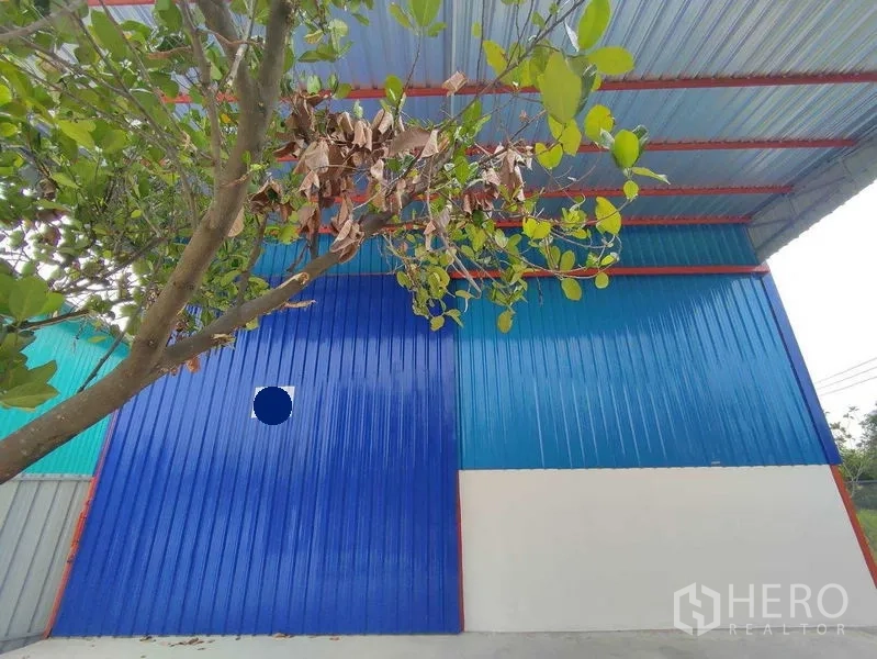 Warehouse for rent in Pak Kret, Nonthaburi - Close-up of blue metal-clad warehouse facade shaded by a tree.