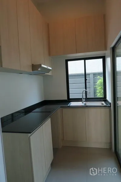 Townhouse for rent in Suan Luang, Bangkok - Compact L-shaped kitchen with light wood cabinets, black countertop, window, and sink.