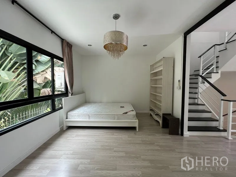 Townhouse for sale in Bang Kapi, Bangkok - Ground-floor bedroom with bed, shelving and large window beside staircase.