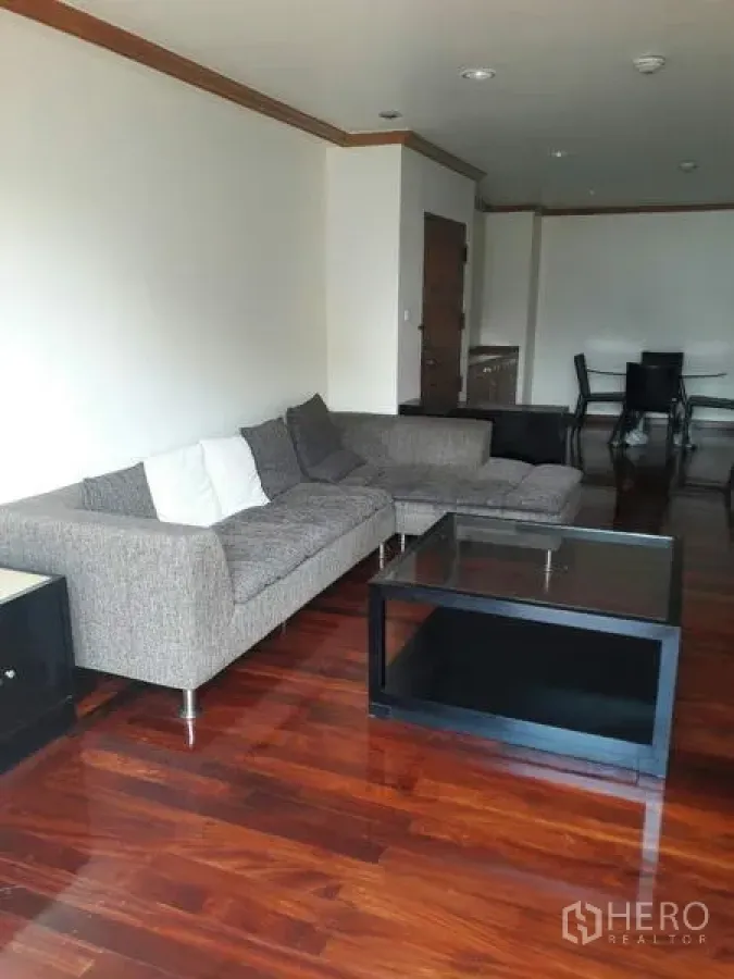 Condominium for rent in Pathum Wan, Bangkok - Cozy living room corner featuring a gray L‑shaped sofa and glass coffee table on gleaming wooden floors.