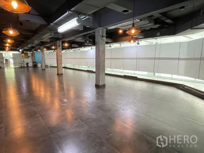 Office Space for rent in Ratchathewi, Bangkok - Spacious open-plan office floor with dark tiles, exposed ceiling, pendant lights, and a long wall of shaded windows.