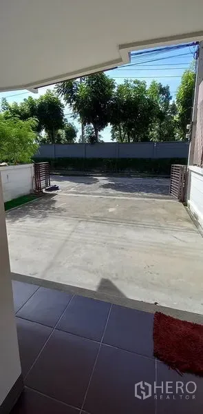 Townhouse for sale in Mueang Pathum Thani, Pathum Thani - Covered carport and wide concrete driveway behind a sliding gate with trees beyond.