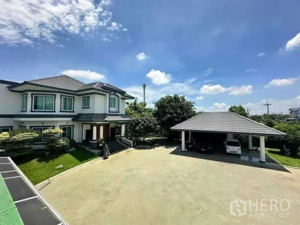 Detached House for sale in Khlong Luang, Pathum Thani - Expansive driveway with the main two-storey house and a covered carport amid green lawns under a blue sky.