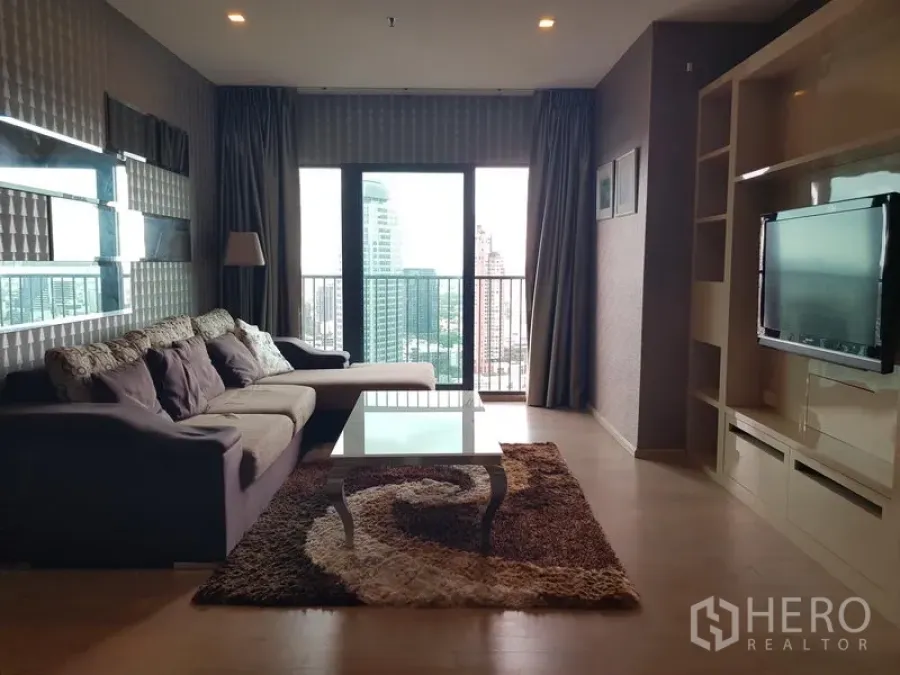 Condominium for rent in Khlong Toei, Bangkok - Spacious living room with sectional sofa, TV wall unit and balcony overlooking the Bangkok skyline.