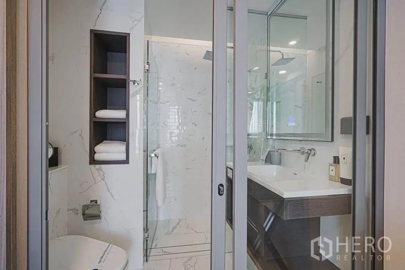 Condominium for sale in Khlong Toei, Bangkok - Modern bathroom featuring glass shower, white marble tiles, large mirror and storage shelves.
