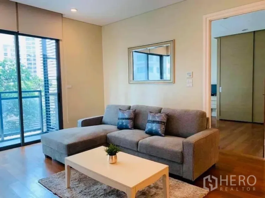 Condominium for rent in Khlong Toei, Bangkok - Bright Sukhumvit 24 living room with a grey sectional sofa, coffee table, and balcony overlooking city greenery.