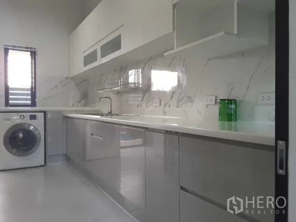 Detached House for rent or sale in Lam Luk Ka, Pathum Thani - Bright white kitchen with marble backsplash, long countertop, upper cabinets and a washing machine by the window.