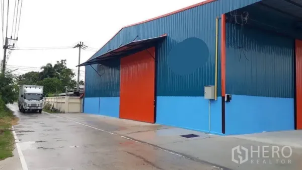 Factory for rent in Khlong Luang, Pathum Thani - Street-side view of a factory bay with a large orange door and truck access along a concrete road.