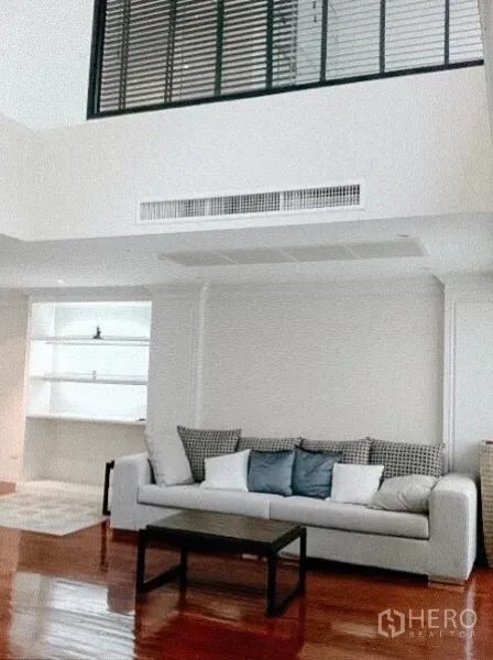 Townhouse for rent in Khlong Toei, Bangkok - Spacious living room with high ceiling and mezzanine-style window, sofa seating and built‑in shelves.