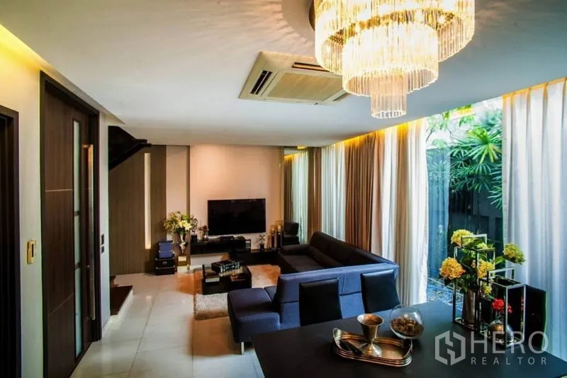 Townhouse for rent or sale in Watthana, Bangkok - Open-plan living room with sectional sofa, chandelier and floor-to-ceiling windows facing a small garden