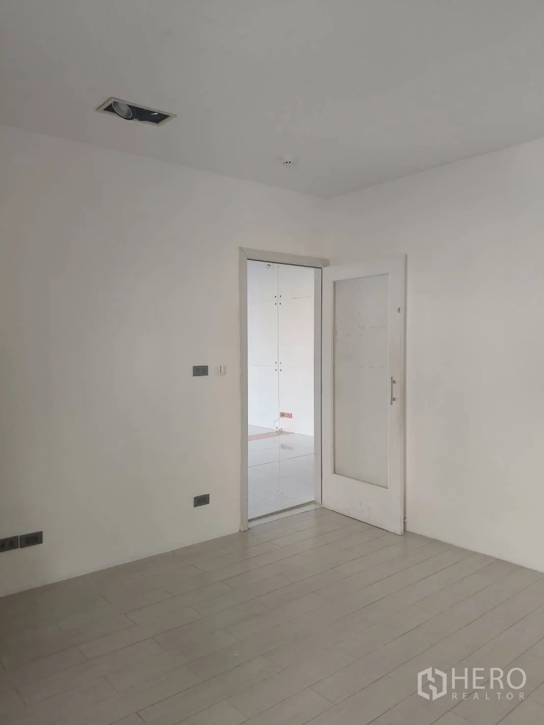 Office Space for rent in Khlong Toei, Bangkok - Minimal white room with light wood flooring and a glass door opening to the adjoining tiled office area.