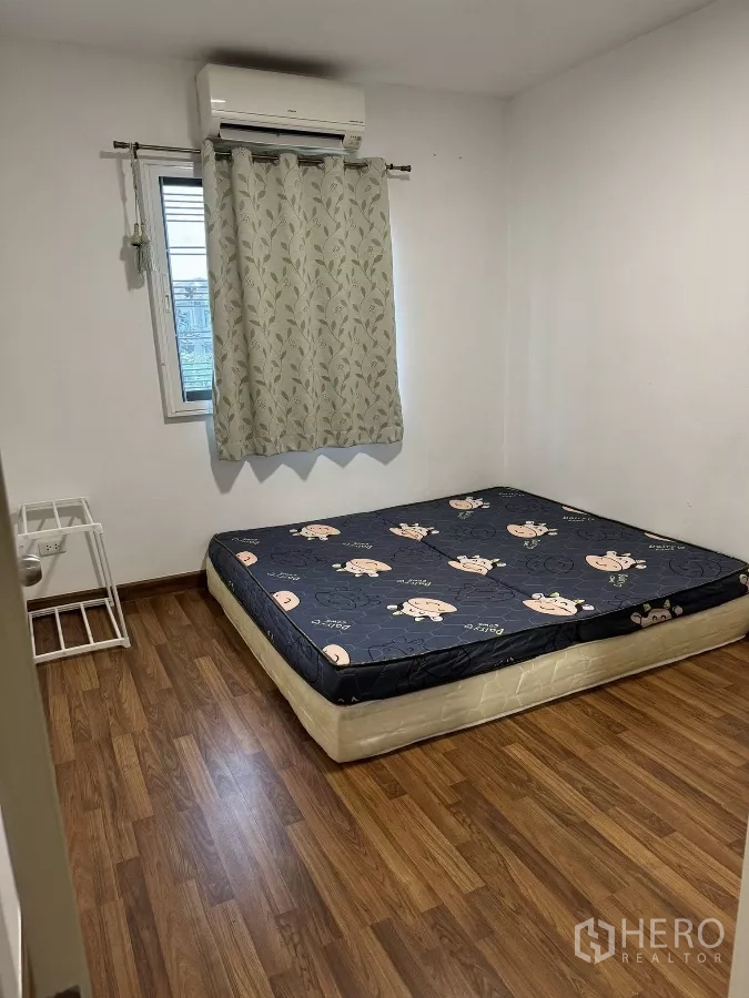 Townhouse for rent in Suan Luang, Bangkok - Simple bedroom with wood-look flooring, window with curtains, and wall-mounted air conditioner.