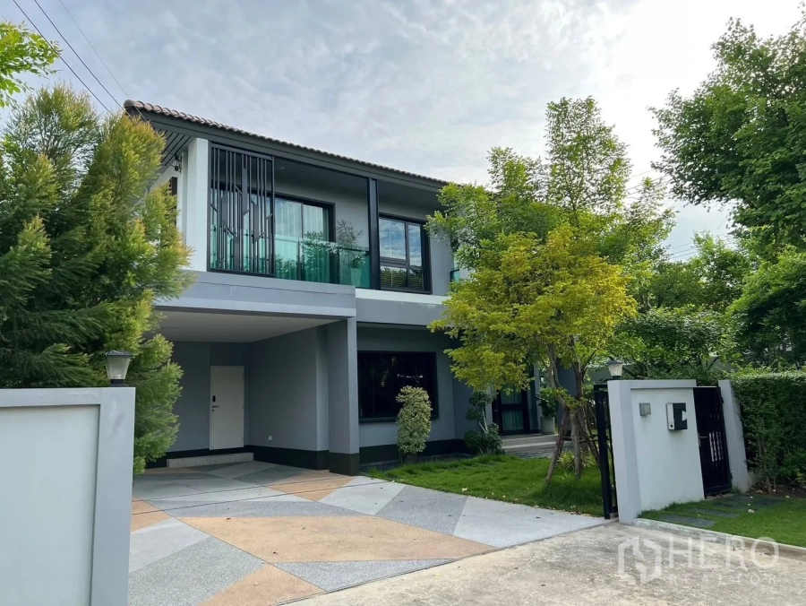 Detached House for rent in Bang Phli, Samut Prakan - Front facade with driveway, covered carport, lush trees, and a gated entrance.