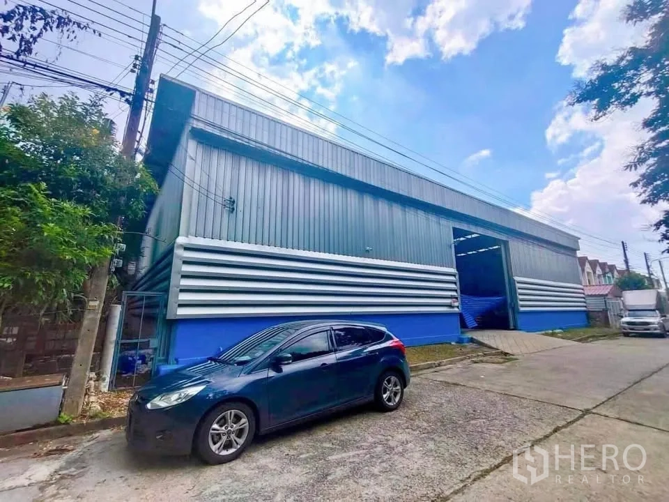 Warehouse for rent in Thanyaburi, Pathum Thani - Front exterior of the warehouse with open loading door, blue base cladding, and driveway access for vehicles.