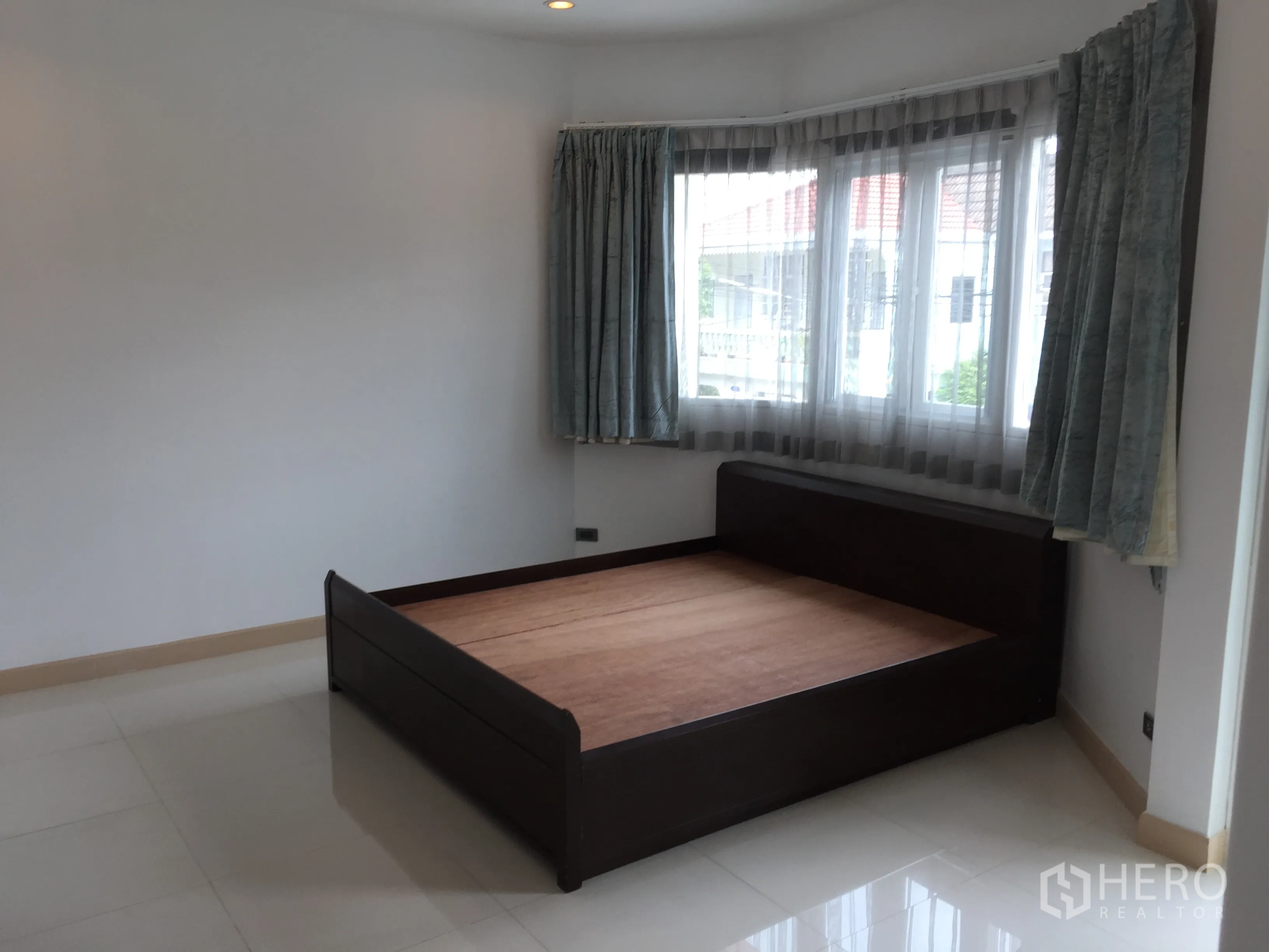 Townhouse for rent in Din Daeng, Bangkok - Bright bedroom with corner windows, sheer curtains and a dark wooden bed frame on glossy tile floors.