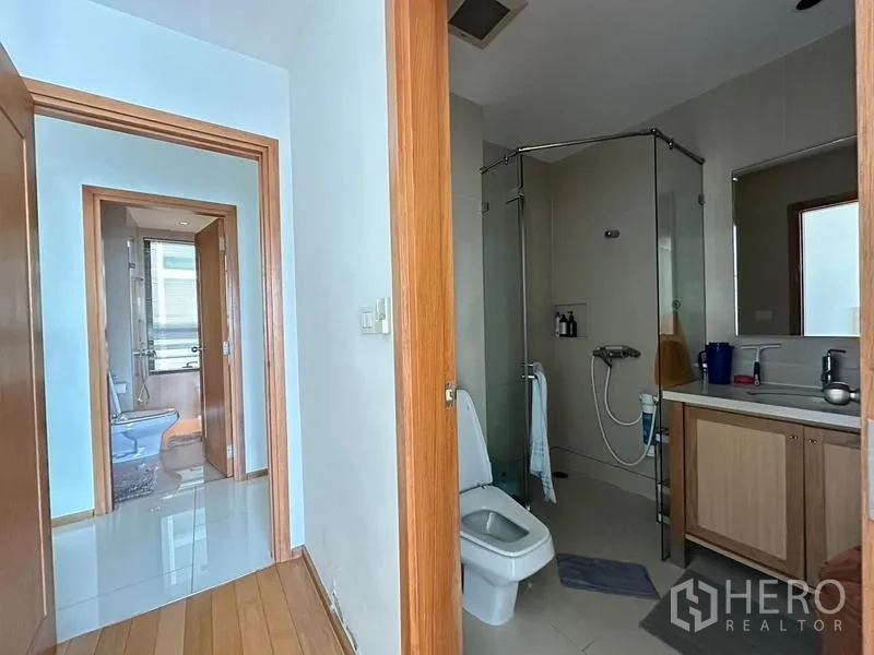 Condominium for sale in Khlong Toei, Bangkok - Bathroom with glass shower, toilet and vanity; hallway leading to an additional bath.