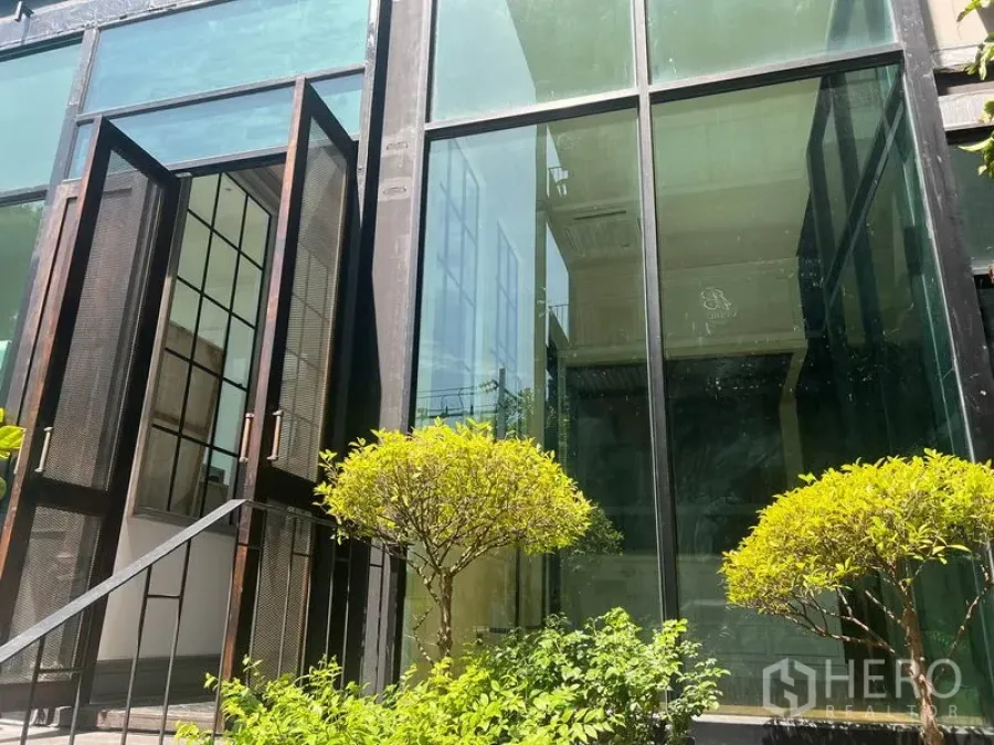 Retail Space for rent in Khlong Toei, Bangkok - Street-facing glass storefront with open steel-framed doors and trimmed shrubs at the entrance of a Thonglor retail unit.