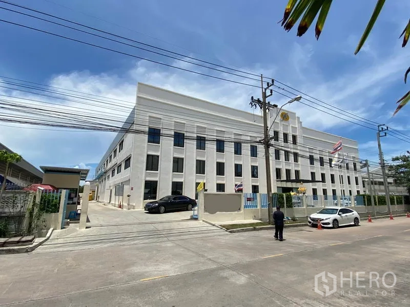 Factory for rent or sale in Bang Sao Thong, Samut Prakan - Street view of a modern multi-storey factory compound with front gate, parking area, perimeter fence and overhead power lines on a sunny day.