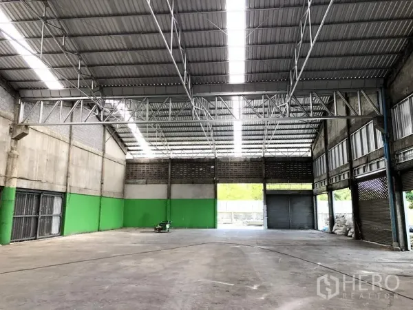 Warehouse for rent in Pathum Thani - Spacious warehouse interior with high steel truss roof, skylight panels, concrete floor, and large roller shutter doors.