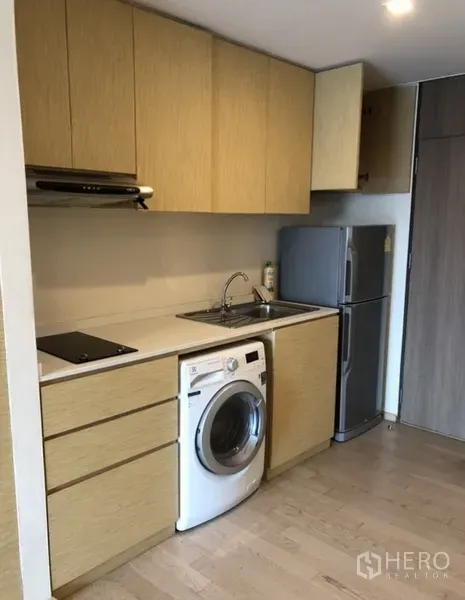 Condominium for rent in Khlong Toei, Bangkok - Compact kitchen with light wood cabinets, sink, electric cooktop, washing machine and refrigerator.