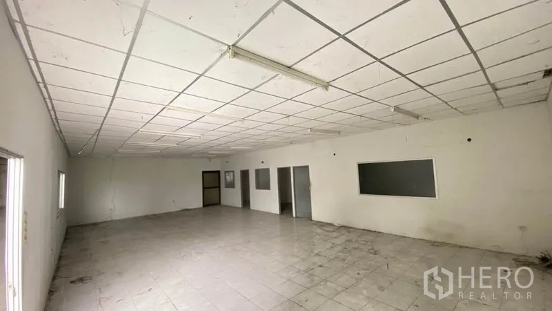 Warehouse for rent in Samut Prakan - Empty office room with tiled floor, drop ceiling, fluorescent lights and interior windows.
