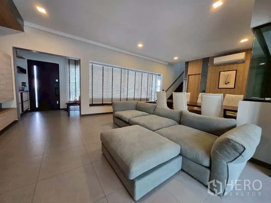 Townhouse for rent in Bang Phli, Samut Prakan - Spacious living area with sectional sofa, floor-to-ceiling blinds, recessed lighting, and the entryway in view.