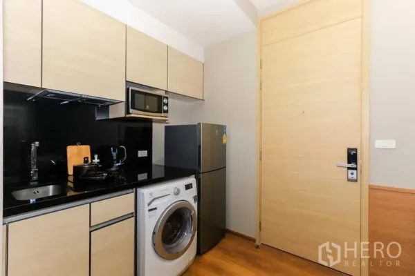 Condominium for rent in Khlong Toei, Bangkok - Compact modern kitchenette with black backsplash, microwave, fridge and front-load washing machine.