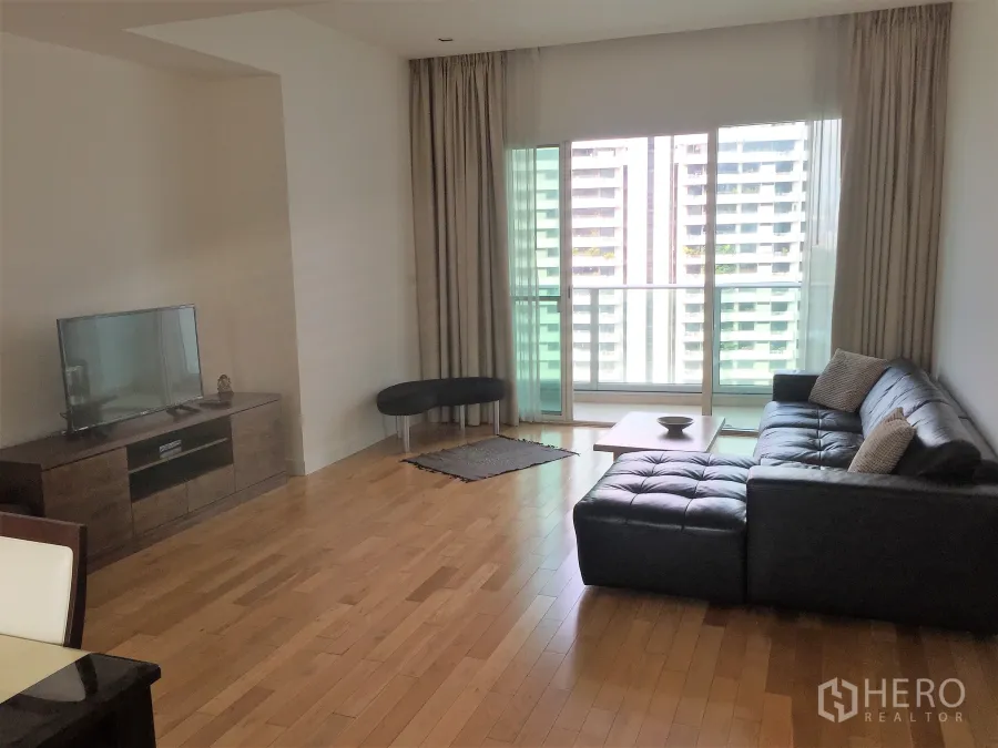Condominium for rent in Khlong Toei, Bangkok - Bright living room with black leather sectional, wood floors, TV console, and sliding doors to a balcony with city view.